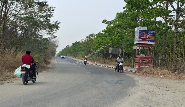 Motorcyclists traveling along a lush green paved road with an advertising billboard, showcasing the well-connected location of Kollur, Hyderabad.