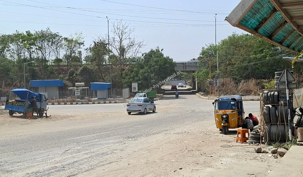A street junction featuring auto-rickshaws, small stalls, and a railway overbridge in the distance, demonstrating the local transit points at the location of Kollur, Hyderabad.